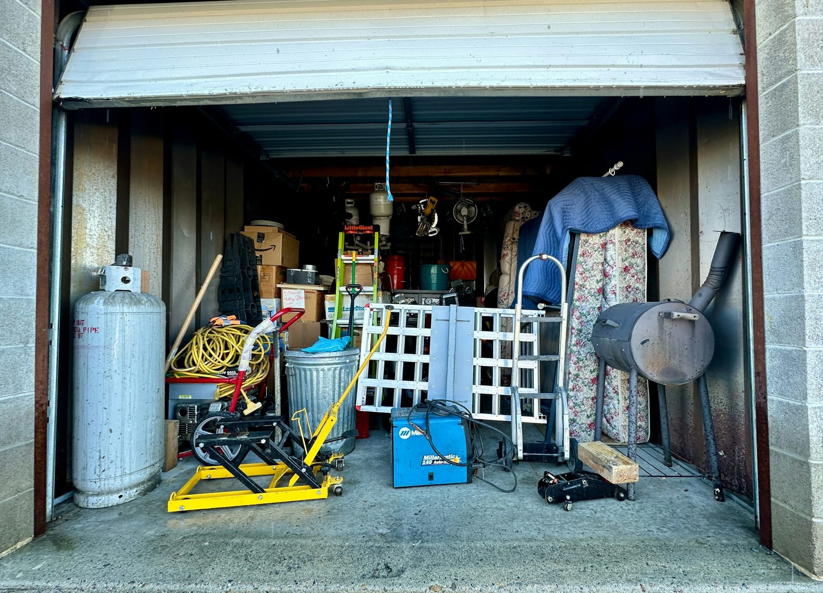 A packed single-car garage: propane tank, extension cord, ladder, hand truck, welder, mattress and a barrel smoker.